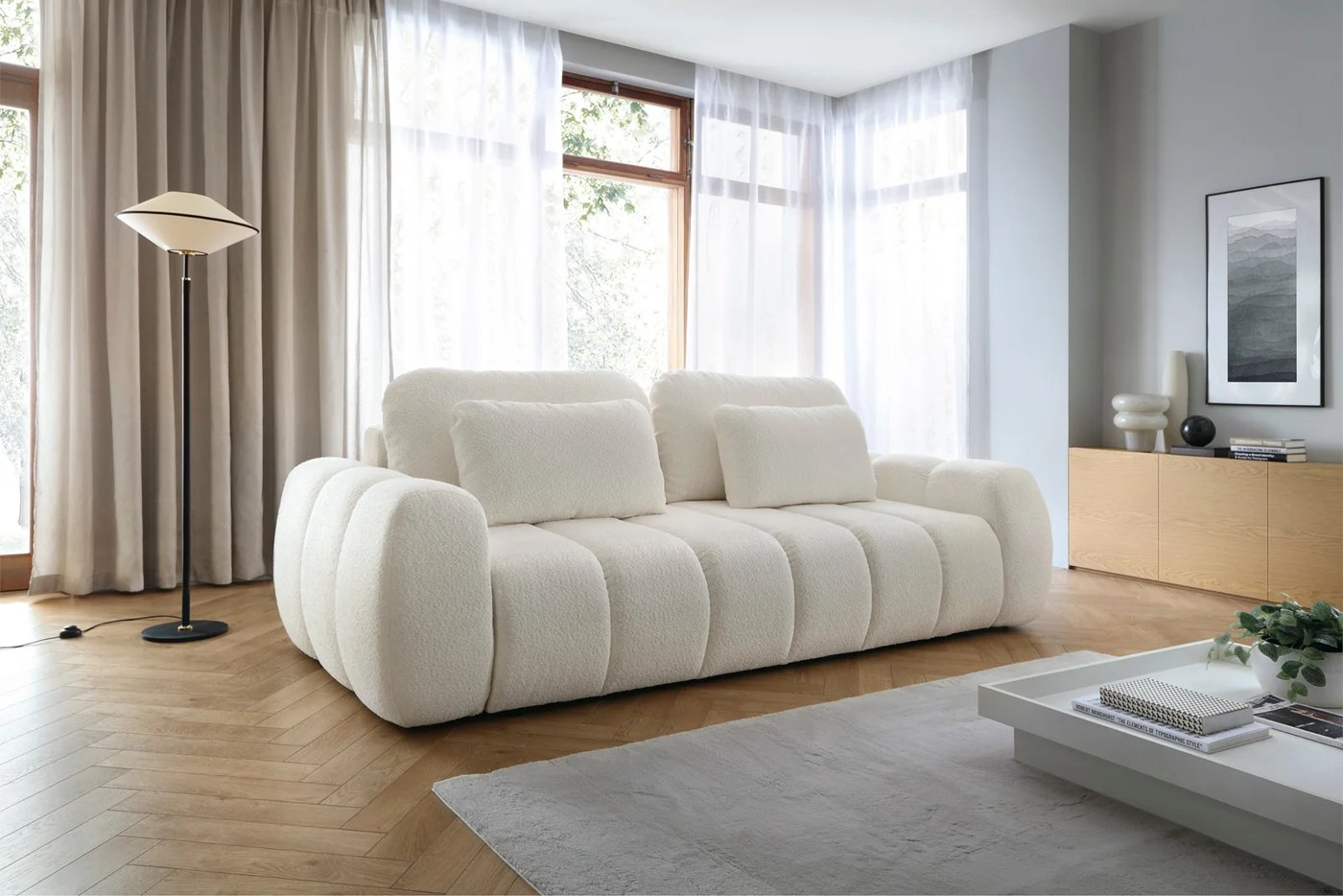 Svetlá pohovka DERBY SOFA so zaoblenými podrúčkami a opierkovými vankúšmi v interiéri pri okne.